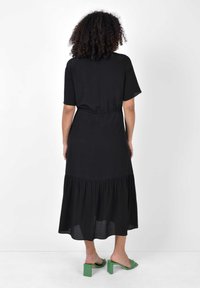 Svart, ankellång klänning med korta ärmar, elastisk midja och lager på lager-skirt. Stylad med gröna klacksandaler. Slät textur.