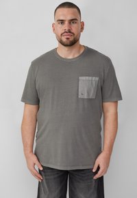 Szara koszulka z krótkim rękawem, z kieszonką na piersi w jaśniejszym szarym odcieniu. Tkanina wydaje się być miękka i lekko teksturowana.