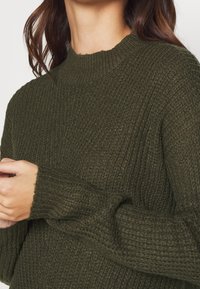 Maglione lavorato a maglia verde scuro con un collo alto, tessuto strutturato e polsini a coste, che mette in mostra un design accogliente e informale.