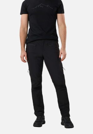 Mężczyzna w czarnej koszulce z motywem górskim, czarnych spodniach typu cargo z kieszeniami na zamek oraz czarnych butach trekkingowych, stojący na białym tle.