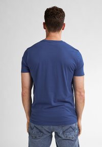 T-shirt blu a maniche corte con scollo rotondo; tessuto di cotone liscio; design aderente; indossato con jeans in denim azzurro, visto da dietro.