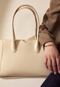 Sac fourre-tout en cuir beige avec deux poignées, surface texturée, détail en V en haut et logo doré sur le devant. Accents dorés sur une main.