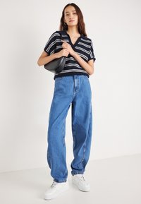 Navy μπλε ριγέ πουλόβερ κοντομάνικο με V λαιμόκοψη, συνδυασμένο με φαρδιά μπλε τζιν παντελόνια και λευκά αθλητικά παπούτσια. Μια μαύρη τσάντα ώμου ολοκληρώνει την εμφάνιση.