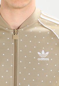 Beige zip-up jacket made of fabric with white triangle patterns. Features white Adidas logo and three white stripes on the shoulders.