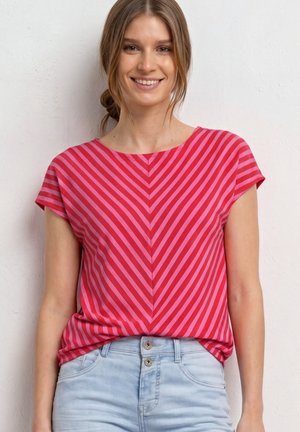 Žena se usmívá, oblečená do červeného a růžového pruhovaného trička s krátkým rukávem zastrčeného do světle modrých džínů s vysokým pasem, stojící před jednoduchou zdí.