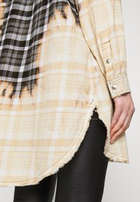 Chemise à manches longues à carreaux beige et noir avec ourlet effiloché portée sur un legging noir, montrant une fente latérale et un poignet avec des boutons-pression métalliques.