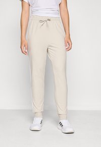 Bežové tepláky s elastickým pásom a nastaviteľným šnúrkou, s bielymi bočnými pruhmi a tesným manžetou na členkoch.