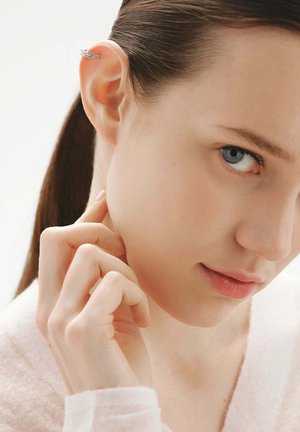 Mujer joven de piel pálida y ojos azules que lleva un brazalete de plata en la oreja, tocándose el costado del cuello con la mano.