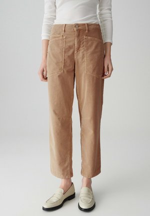 Beige Cordhosen mit einem lockeren Schnitt, ausgestattet mit zwei großen Fronttaschen und einem Metallknopfverschluss, kombiniert mit hellen Slip-On-Schuhen.