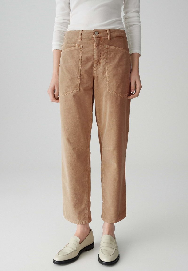 Pantalon en velours côtelé beige avec une coupe décontractée, doté de deux grandes poches avant et d'une fermeture à bouton en métal, associé à des chaussures à enfiler de couleur claire.