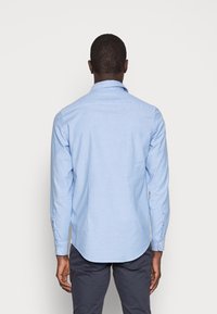 Hombre de pie de espaldas, vistiendo una camisa de botones de manga larga azul claro y pantalones gris oscuro, contra un fondo blanco liso.