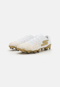Des crampons de football blancs avec une tige texturée, présentant un motif craqué doré sur les orteils et une semelle dorée avec des pointes noires.