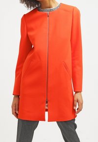 Jasně oranžový kabát s kulatým výstřihem, plným zipem a bočními kapsami, se strukturální šedou stahovací límcem. Hladký povrch tkaniny.