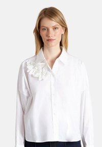 Camisa blanca de manga larga con un frente abotonado y un acento de apliqué floral tridimensional en el hombro izquierdo. Textura de algodón suave.
