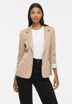 Femme aux longs cheveux foncés portant un blazer beige par-dessus une chemise blanche et un pantalon noir, debout devant un fond clair uni.