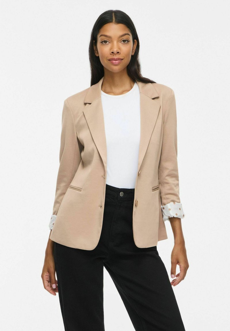 Femme aux longs cheveux foncés portant un blazer beige par-dessus une chemise blanche et un pantalon noir, debout devant un fond clair uni.