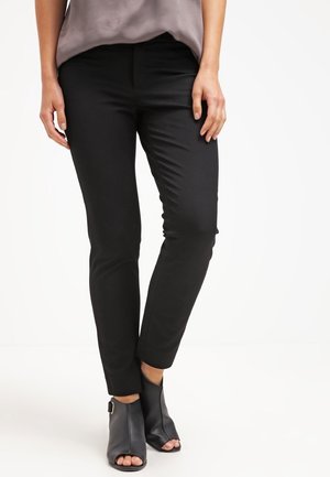 Vrouw draagt nauwsluitende zwarte broek met enkelhoge zoom en zwarte open-toe enkellaarsjes met hak, staand tegen een witte achtergrond.