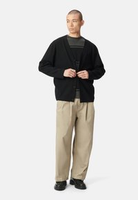 Cárdigan negro con botones, textura suave, llevado sobre una camisa a rayas gris. Combinado con pantalones beige de corte holgado y botas negras gruesas.