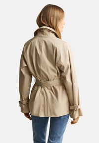 Γυναίκα με μπεζ καμπαρντίνα με ζώνη και αγκράφες στα μανίκια, από πίσω, συνδυασμένη με μπλε τζιν.