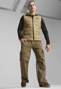 Gilet trapuntato beige con colletto alto e zip frontale, abbinato a pantaloni cargo oliva con più tasche e una vestibilità comoda. Scarpe marroni.