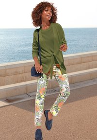 Groene gebreide cardigan over een groene top, bloemenpatroon broek in felle kleuren, en blauwe instappers, aangevuld met een kleine marineblauwe tas.