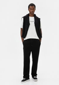 Chaleco sin mangas de forro polar negro sobre una camiseta blanca con texto negro, combinado con pantalones jogger negros y zapatillas negras con suelas blancas.