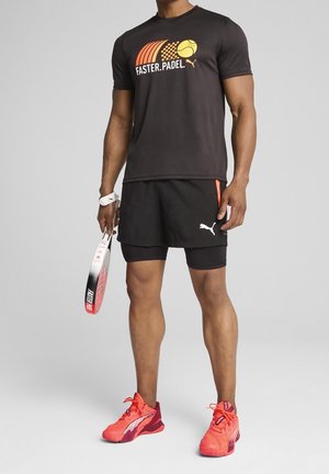 Uomo con maglietta nera "Faster.Padel" e pantaloncini, che tiene una racchetta da padel, indossa scarpe sportive arancioni brillanti, in piedi su sfondo grigio.