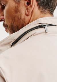 Beżowa kurtka z wysokim kołnierzem, odsłoniętym czarnym zamkiem z tyłu oraz gładką, matową fakturą materiału. Minimalistyczny design z prostymi liniami.