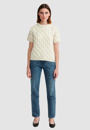 Maglione a maniche corte color crema con un design a trecce, abbinato a jeans blu a gamba dritta e ballerine nere.