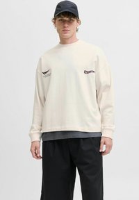 Langærmet cremet sweatshirt med trykte grafikker, lagd over en mørkegrå skjorte, parret med sorte løse shorts og en sort cap.