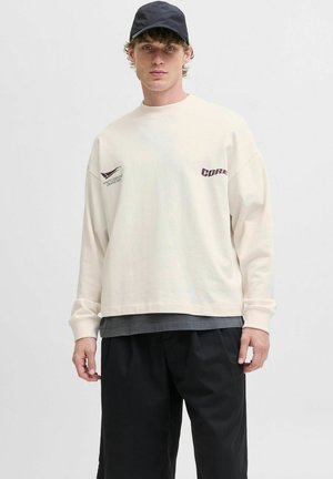Langærmet cremet sweatshirt med trykte grafikker, lagd over en mørkegrå skjorte, parret med sorte løse shorts og en sort cap.