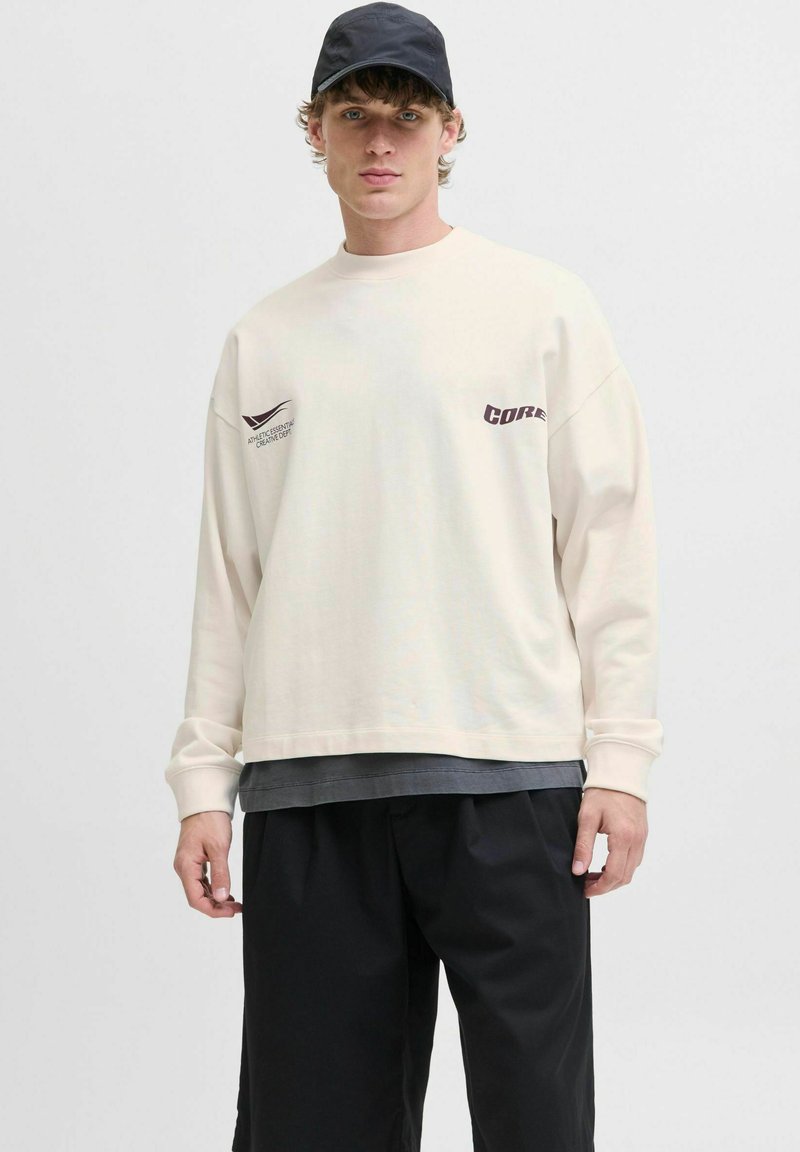 Langærmet cremet sweatshirt med trykte grafikker, lagd over en mørkegrå skjorte, parret med sorte løse shorts og en sort cap.