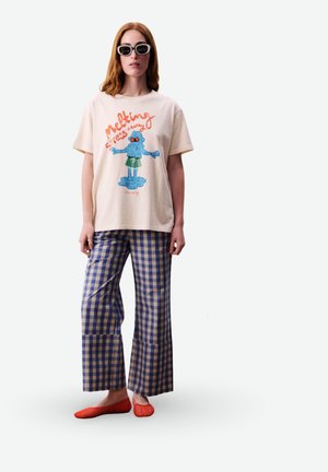 Femme portant un t-shirt graphique blanc avec le texte « melting stress away » et un personnage, pantalon à carreaux bleu, chaussures rouges et lunettes de soleil noires, debout.