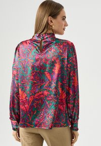 Bluzka z długim rękawem w zielonym satynie z vibrant wzorem paisley w różowym, czerwonym i fioletowym. Posiada dekolt w kształcie łezki oraz falbany przy szyi.