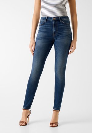 Donna che indossa jeans skinny blu scuro a vita alta abbinati a sandali marroni con cinturini e tacco alto, e una maglietta bianca a righe.