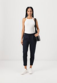 Bílé žebrované tílko, tmavě modré jogger kalhoty s tkaničkou a bílé tenisky; modelka drží černou crossbody tašku. Minimalistický design.
