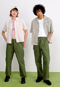 Deux individus portent des tenues décontractées : une chemise à rayures à manches courtes et un pantalon cargo olive, assortis de chaussures foncées. L'arrière-plan est clair.