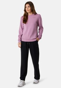 Felpa viola chiaro con scollatura tonda e maniche a sbuffo, abbinata a pantaloni jogger neri con tasche laterali e coulisse, e sneaker bianche.
