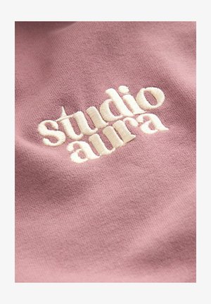 Rožnata tkanina z mehko teksturo, ki prikazuje teksturirano, kremno-beli vezen napis »studio aura«.