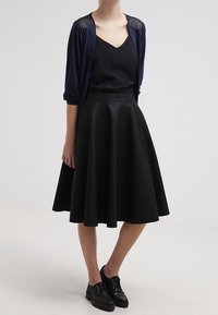 Černá áčková sukně s hladkým povrchem, kombinovaná s námořnickým cardiganem s žebrovanými detaily na ramenou. Černé šněrovací boty doplňují celé oblečení.