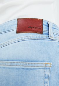 Ljusblå denimjeans med en brun läderetikett som visar "Pepe Jeans London." Har synlig sömnad och flera fickdetaljer.