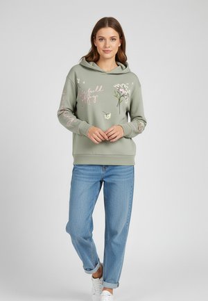 Frau trägt einen hellgrünen Hoodie mit Blumenmustern und dem Text „Leben voller Freude“, kombiniert mit hochgekrempelten blauen Jeans und weißen Sneakern, stehend.