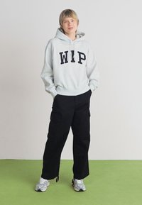 Grijze hoodie met "WIP" in vetgedrukte zwarte letters, gecombineerd met losse zwarte cargobroeken en lichtgrijze sneakers op een groene vloer.