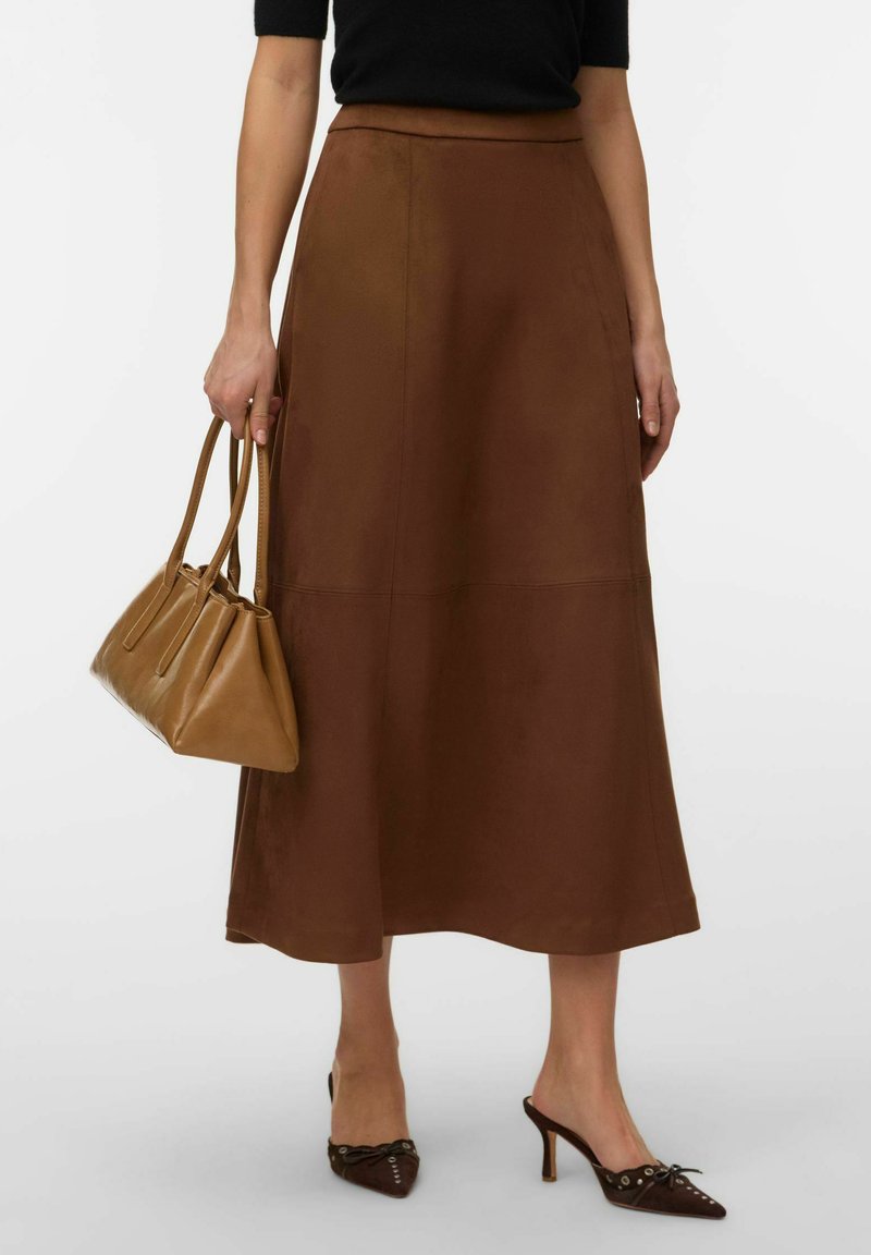Femme portant une jupe midi marron, un haut noir, des chaussures à talons pointues marron, tenant un sac à main en cuir marron clair.