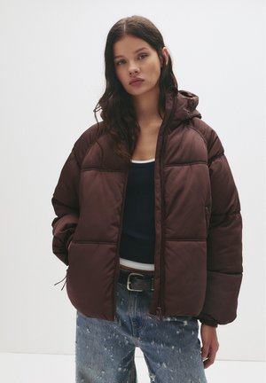 Braune gepolsterte Jacke mit Kapuze, ausgestattet mit einem Reißverschluss und gesteppter Optik. Getragen über einem marineblauen Tanktop und verwaschenen blauen Jeans.