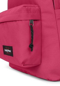 Růžový látkový batoh s přední kapsou, zipovými detaily a černou nášivkou s logem Eastpak. Textura je hladká s vyztuženým švem.