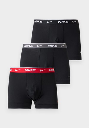Atlétikus férfi mezítláb áll, fekete Nike boxeralsót visel logós derékrésszel, sima fehér háttér előtt.
