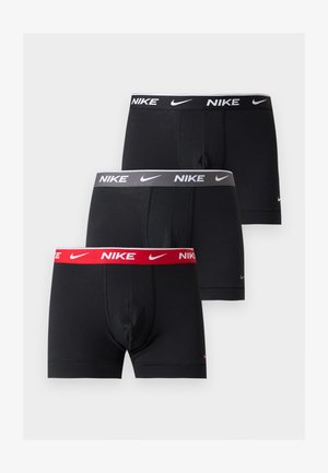 Atletyczny mężczyzna stojący boso, ubrany w czarne bokserki Nike z logo na gumce, na jednolitym białym tle.