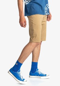 Beige katoenen shorts met voorkeurszakken, gecombineerd met felblauwe canvas sneakers en bijpassende sokken. Het shirt is blauw met een tie-dye patroon.