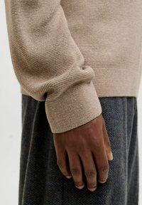 Beige kudumite särk, millel on soonikkuued ja tekstuuriline viimistlus. Käsi restingib tumedal herringbone-mustriga kangal all.